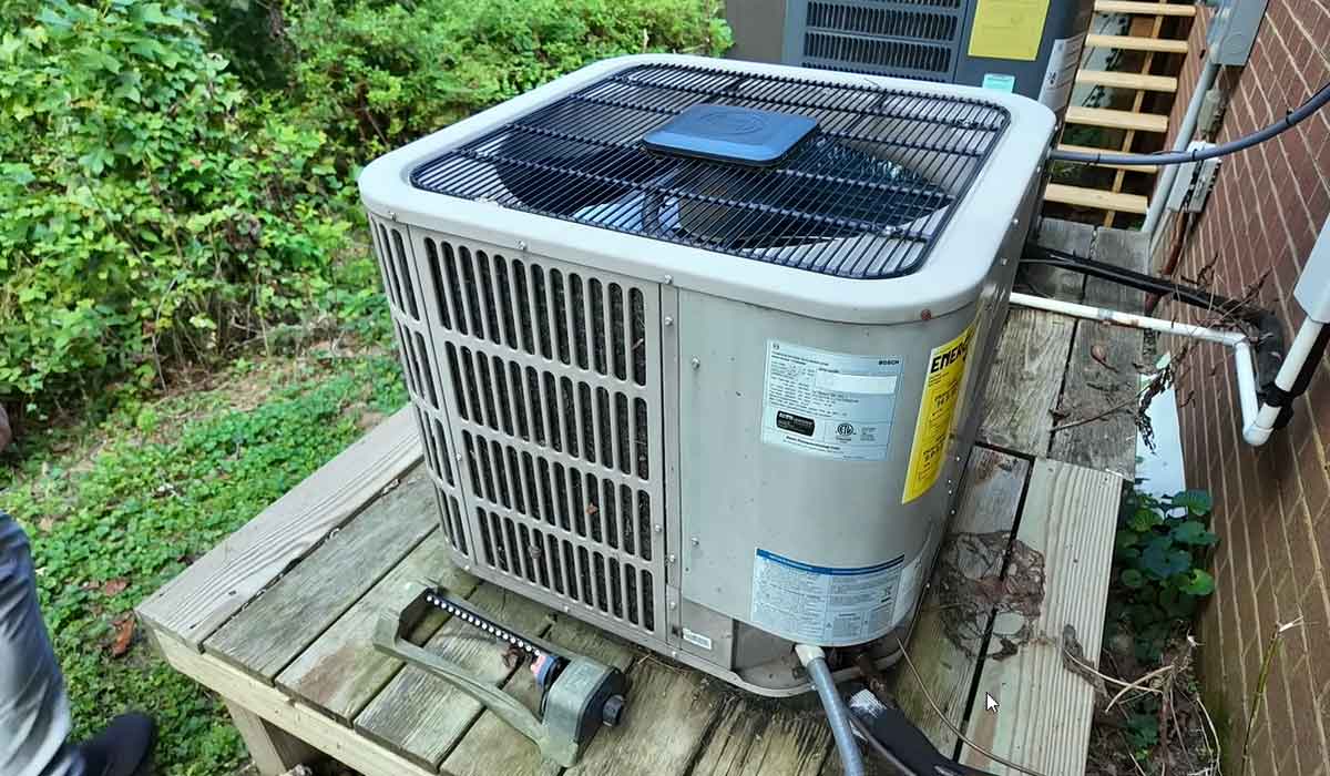 Outdoor AC condenser unit installed on a wooden platform beside a house wall