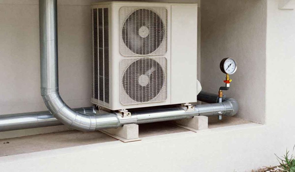 Outdoor HVAC condenser with dual fans, metal piping, and pressure gauge installed beside a building wall