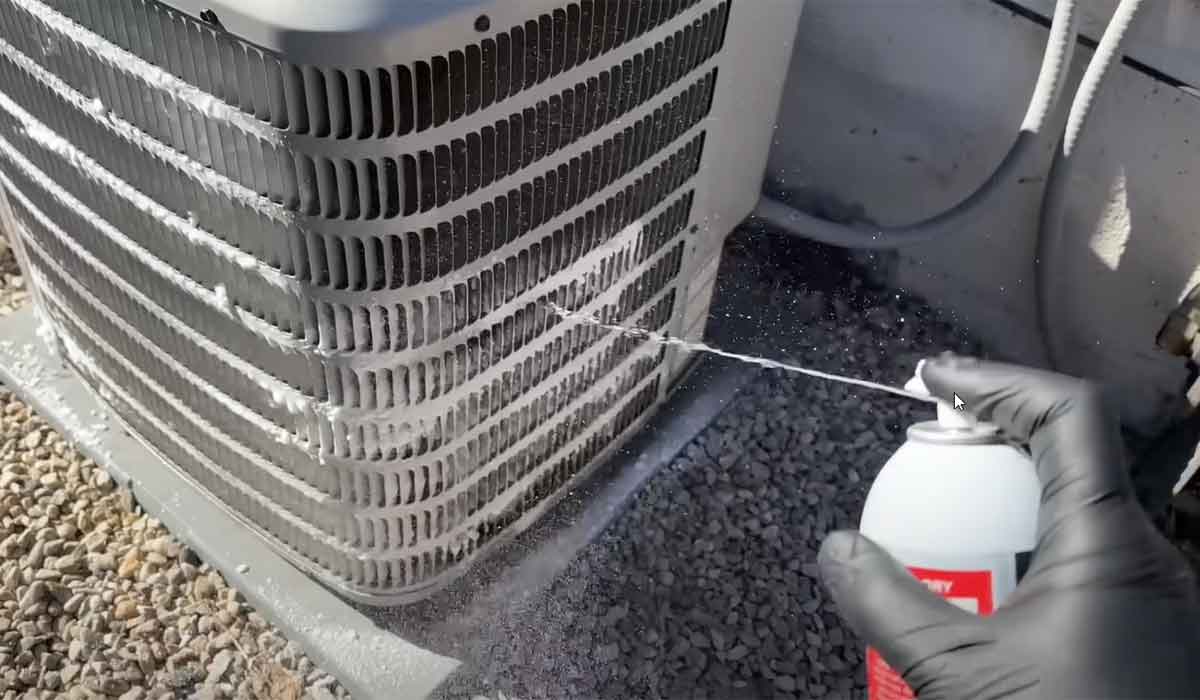 Gloved hand spraying coil cleaner onto an outdoor AC condenser during maintenance