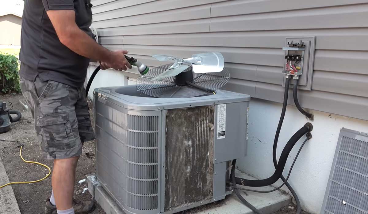 Technician cleaning an outdoor AC condenser with a hose during HVAC maintenance