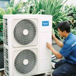 Technician servicing an outdoor HVAC condenser unit beside dense landscaping