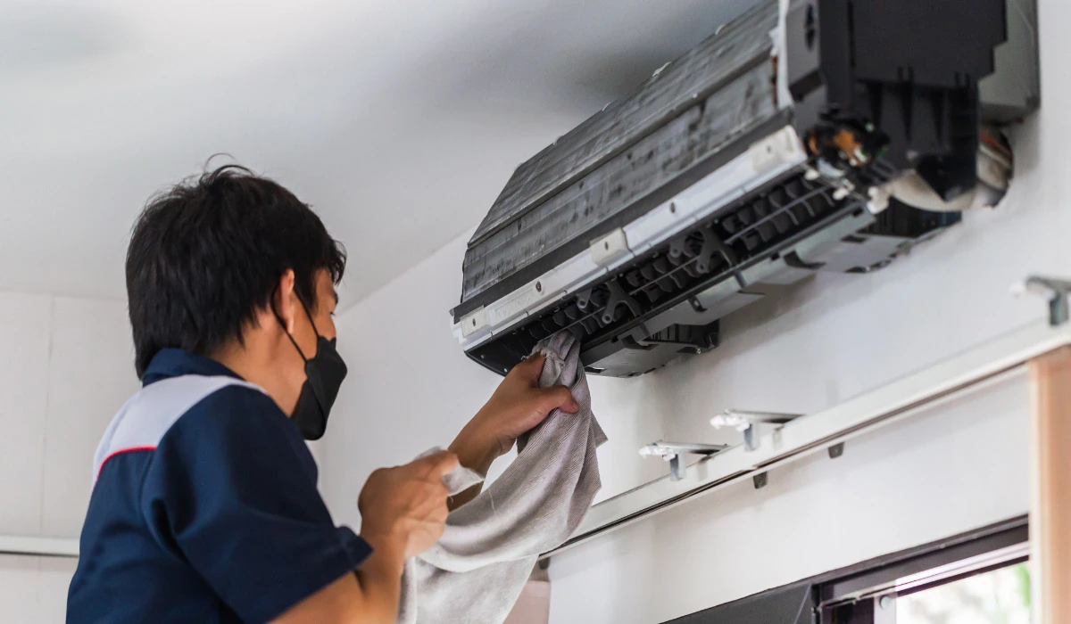 A technician wearing a mask cleans the air conditioning unit mounted on the wall using a cloth to ensure proper maintenance.