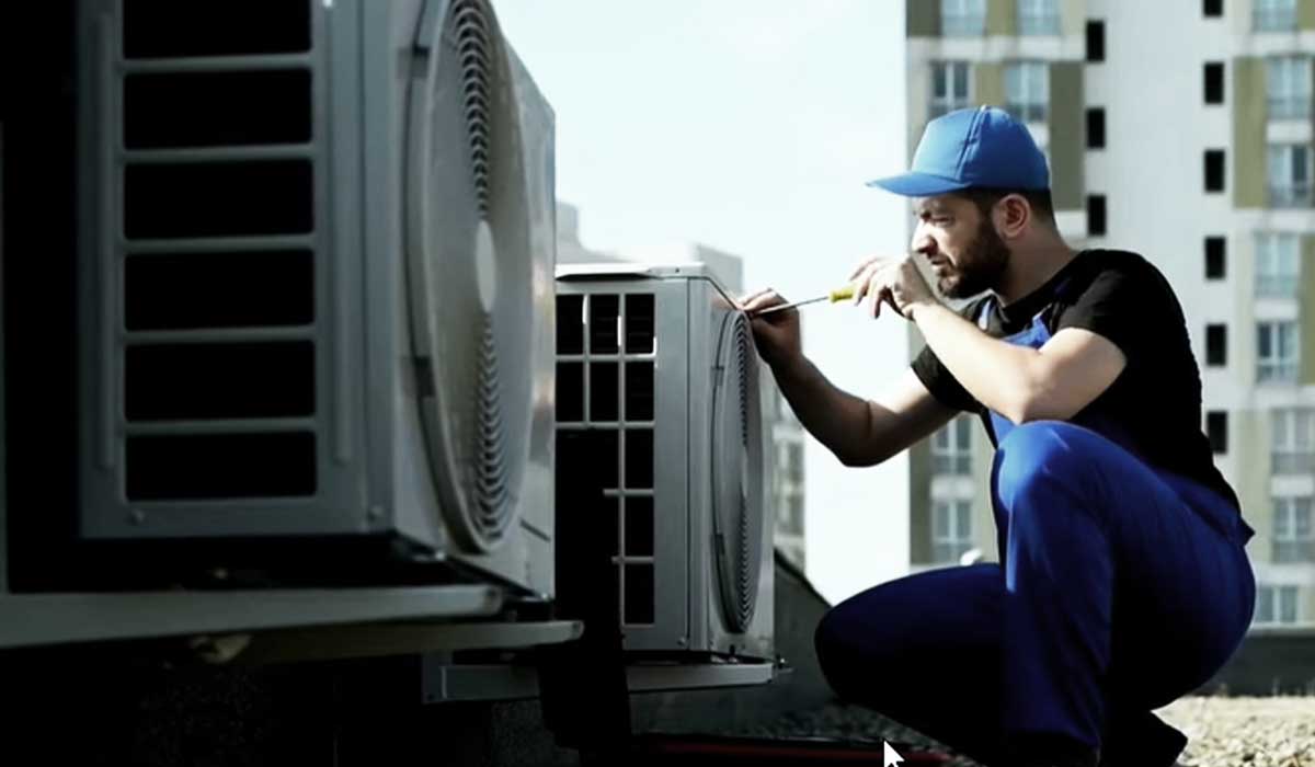 Technician in blue overalls crouching beside AC units, inspecting with a screwdriver.
