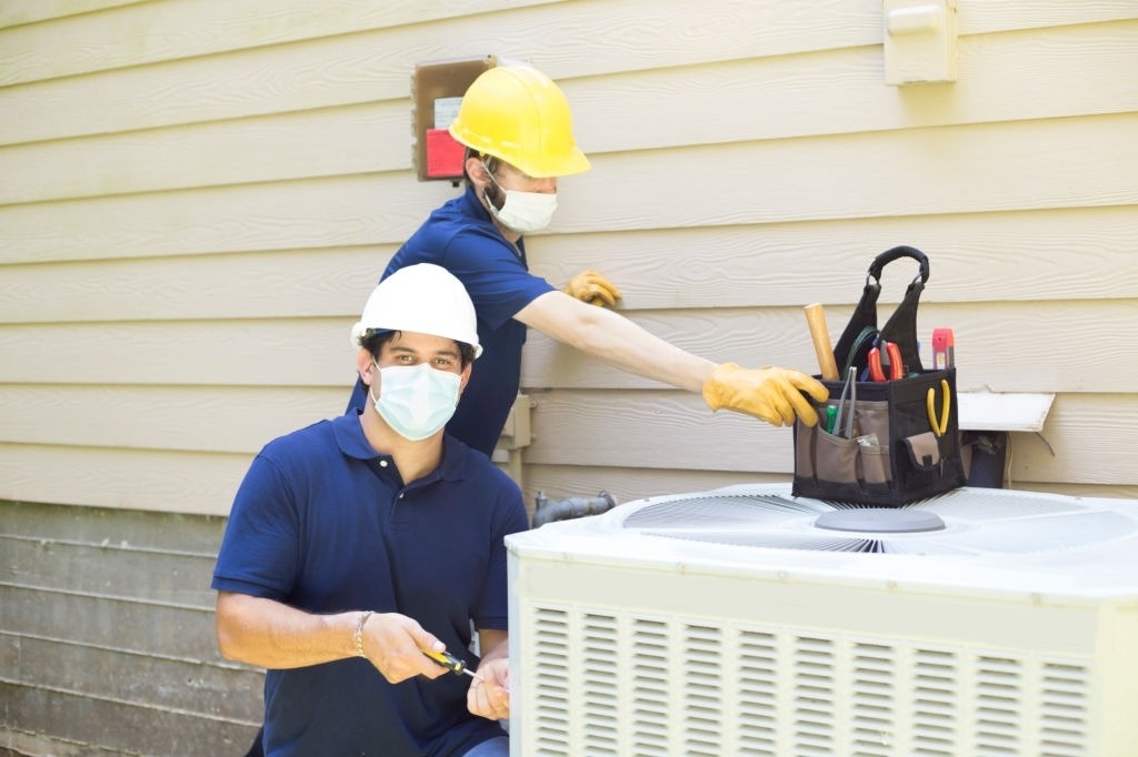 Two workers in safety gear are performing maintenance on an outdoor air conditioning unit, with tools in a belt nearby.