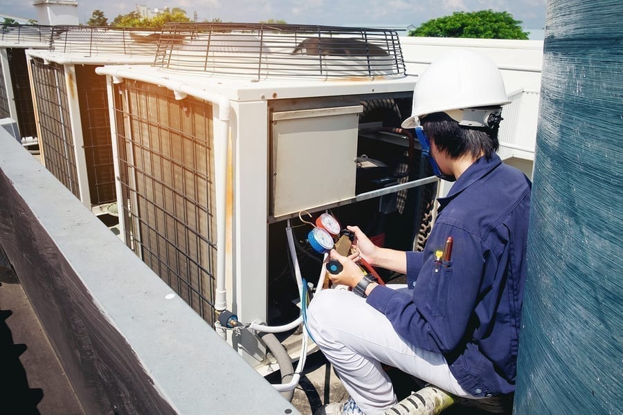 A technician in a hard hat uses pressure gauges to inspect rooftop air conditioning units under a clear sky, representing HVAC services in Greensboro.