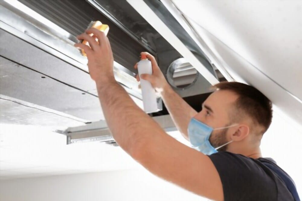 Technician cleaning a ceiling-mounted HVAC vent with spray cleaner and a sponge.