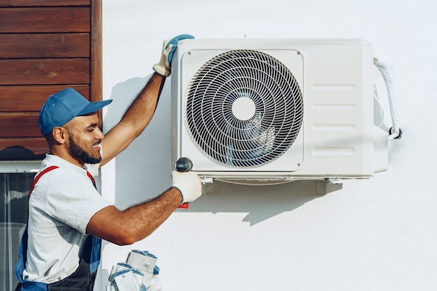 A technician installs an air conditioning unit on a white wall, wearing gloves and a blue cap, with tools at his feet.
