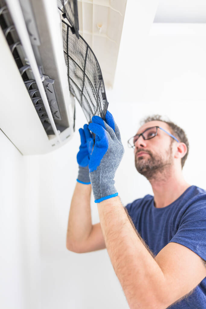 A person wearing gloves removes an air conditioner filter for cleaning in a bright indoor space.