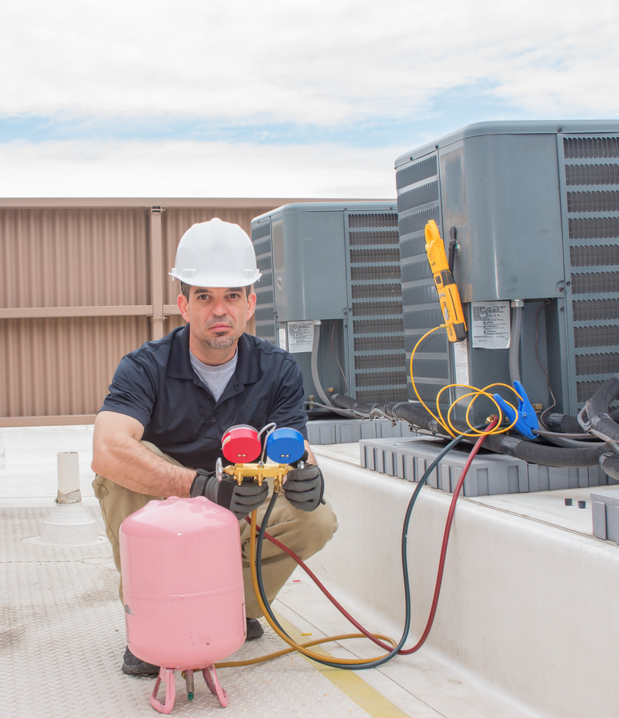 A technician in a hard hat kneels beside AC units in Anna, TX, using a color-coded gauge set and pink refrigerant tank for precise AC repair service.