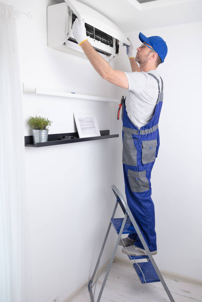 A technician in blue overalls repairs an air conditioner while standing on a stepladder in a bright, modern room.