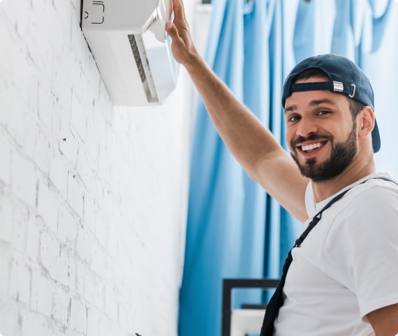 A technician in overalls and cap adjusts a wall-mounted AC unit on a white brick wall, highlighting expert HVAC services in North Carolina homes.