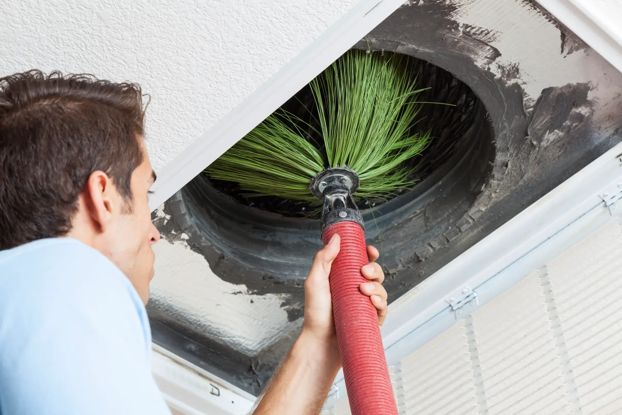 A person uses a red vacuum tool with green bristles to clean a dusty ceiling air duct, showcasing commercial HVAC cleaning and maintenance services.
