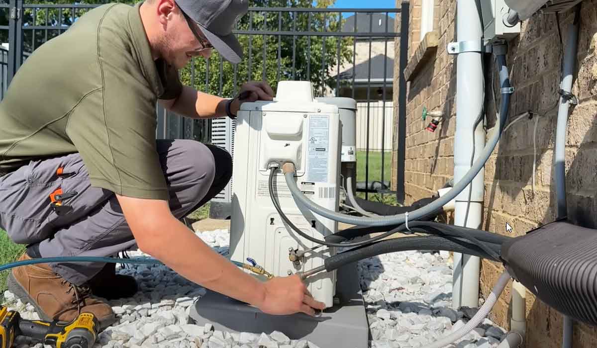A technician in work attire kneels next to an air conditioning unit, connecting pipes and inspecting equipment on a sunny day.