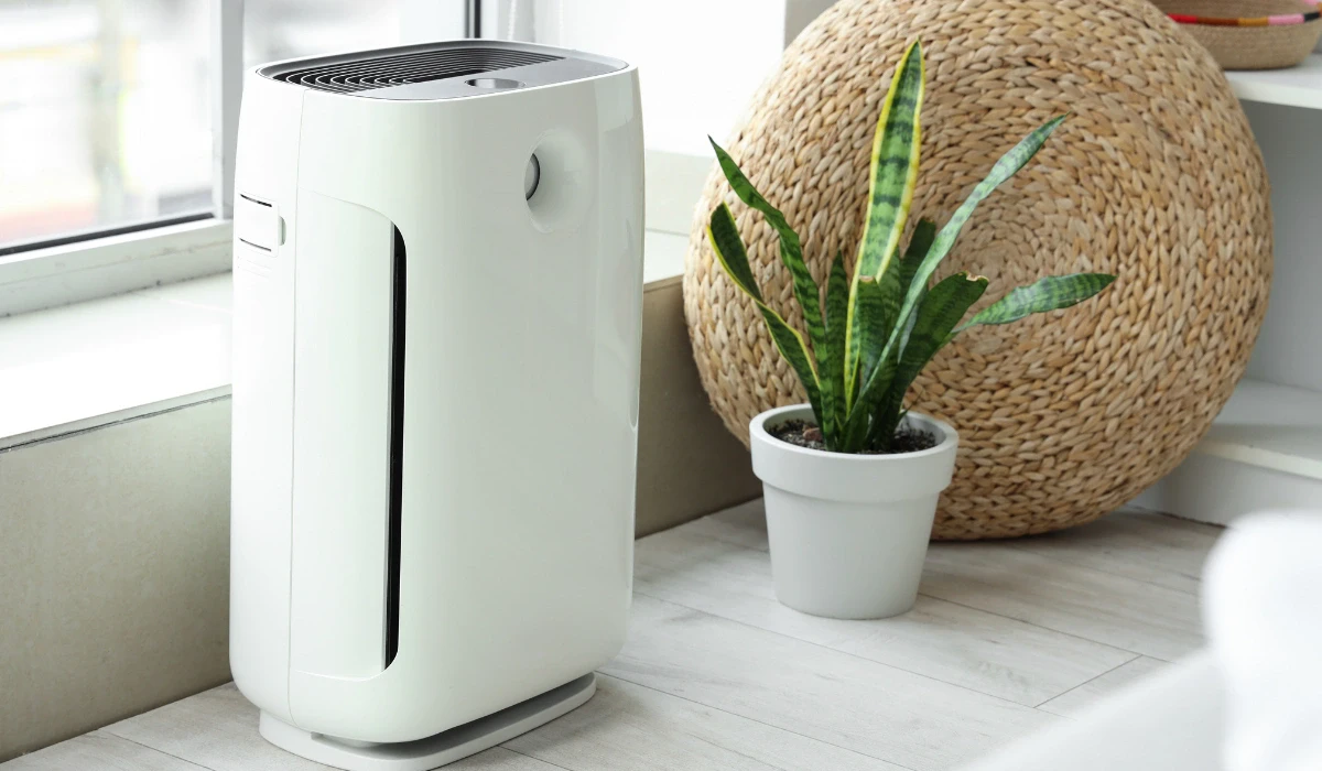 A sleek white air purifier stands on a light hardwood floor beside a potted snake plant and a woven basket, creating a calm, modern atmosphere.